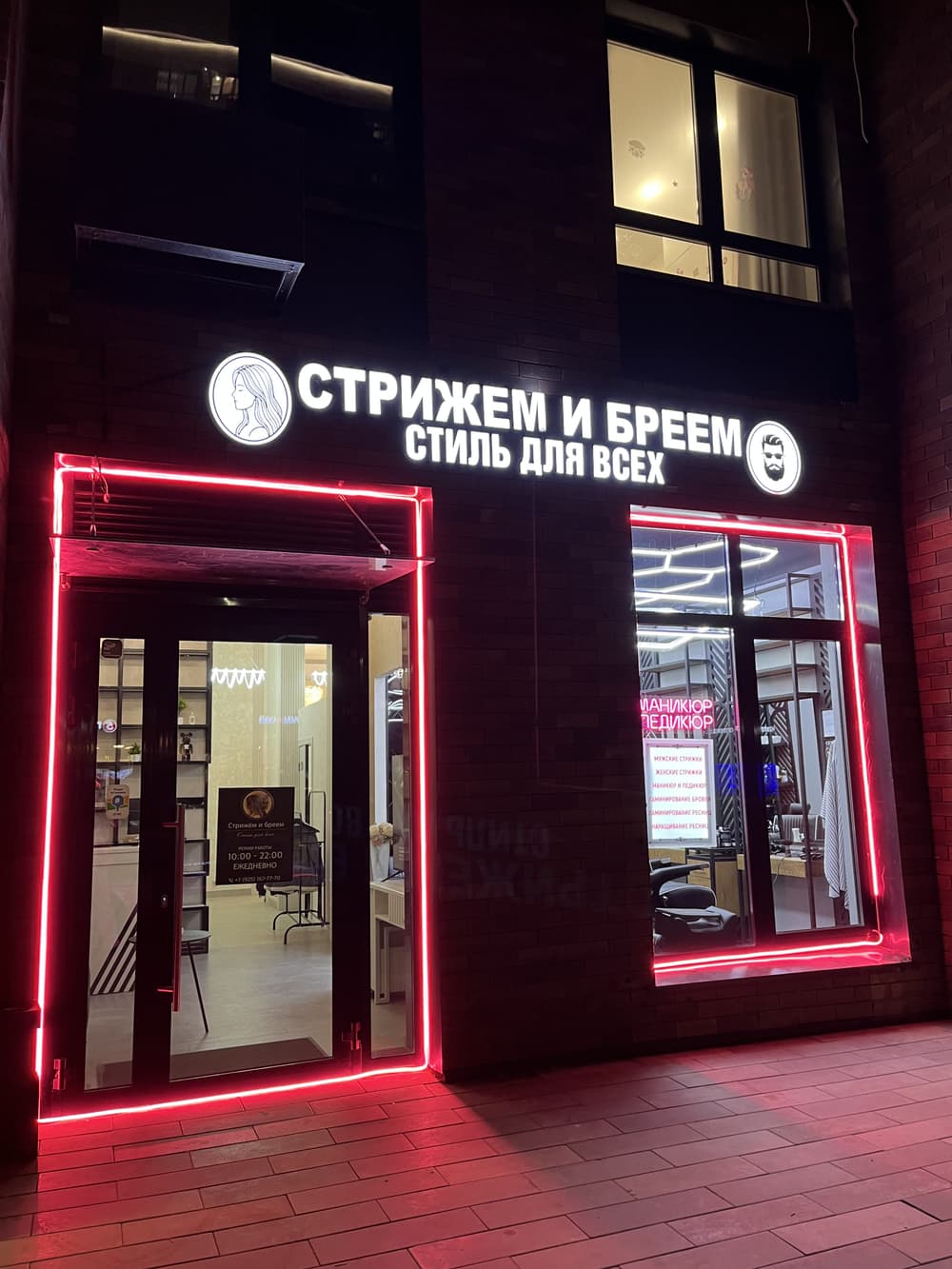Вход в салон вечером: вывеска «Стрижем и бреем» и красная неоновая подсветка двери и окна